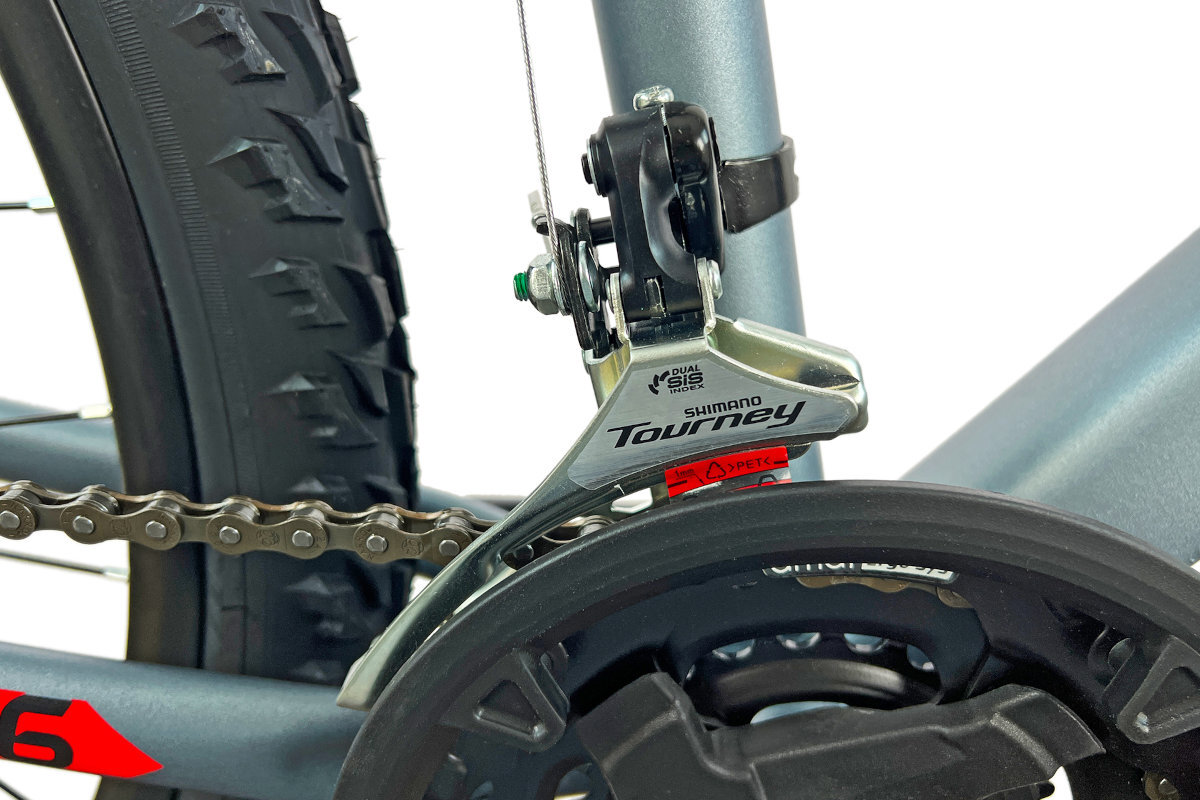 Zbliżenie na mechanizm przerzutki rowerowej z napisem \'Shimano Tourney\'. W tle widoczna jest część opony rowerowej z wyraźnym bieżnikiem.
