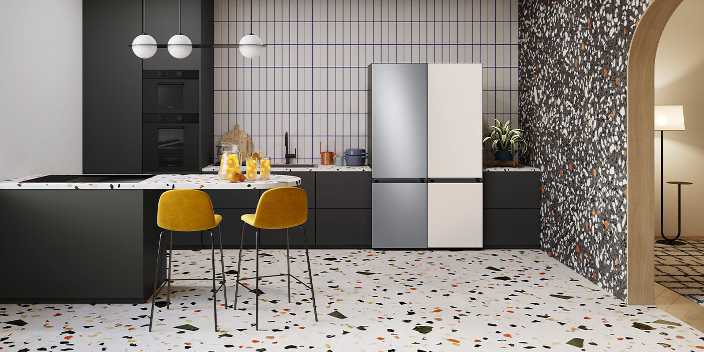 Na ilustracji pokazane są dwie lodówki Samsung z linii BESPOKE, które umieszczone zostały w nowoczesnej kuchni między czarnymi meblami. Lewe urządzenie ma kolor szczotkowanej stali nierdzewnej, a odcień drugiego to kremowy beż.