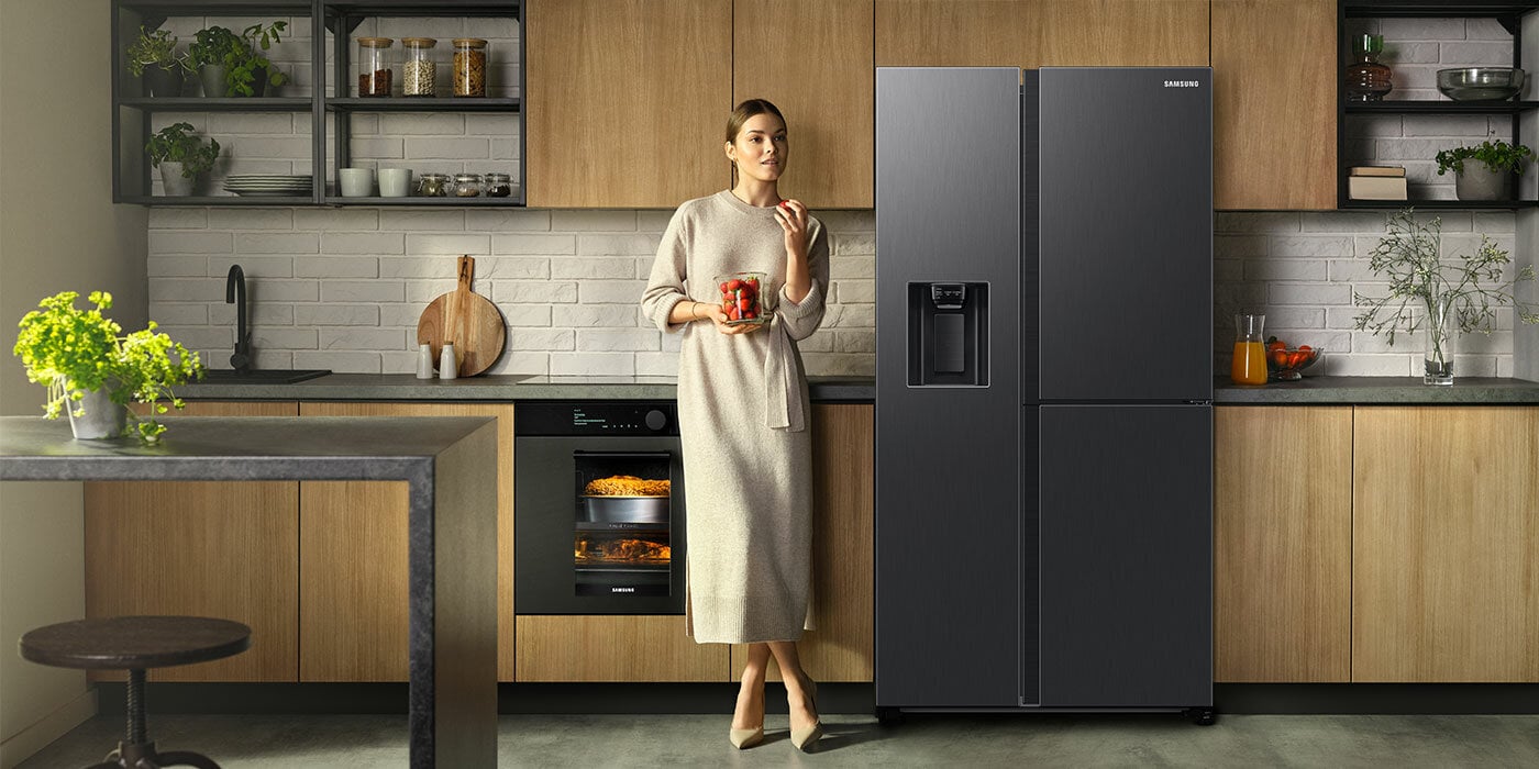 Ilustracja przedstawia użytkowniczkę lodówki Samsung, która stoi zaraz obok urządzenia i trzyma pojemnik z truskawkami.