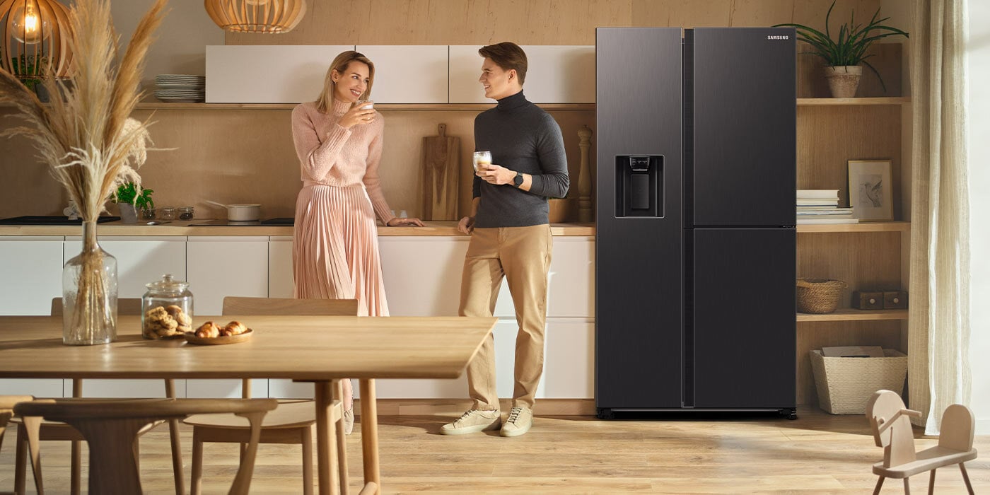 Ilustracja przedstawia kuchnię, w której większość elementów wyposażenia wykonana jest z jasnego drewna. W pomieszczeniu stoi lodówka Samsung w kolorze grafitowej stali, obok której znajdują się jej użytkownicy.
