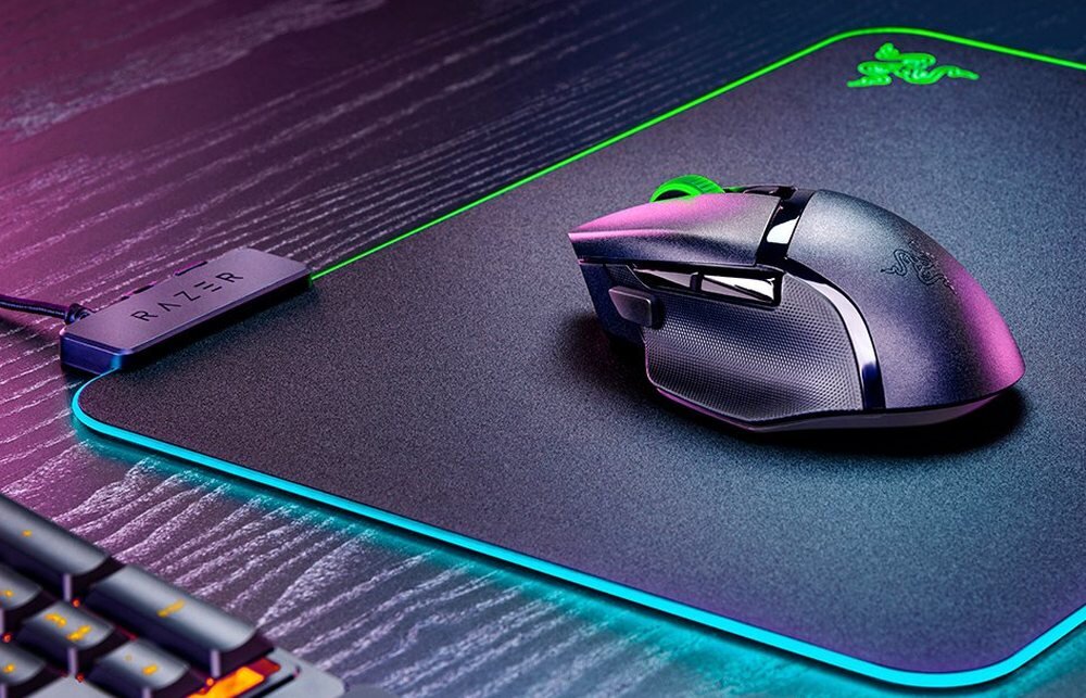 На зображенні зображено чорну комп’ютерну мишу з RGB-підсвічуванням, розміщену на килимку з логотипом RAZER. Поруч із нею знаходиться пристрій із написом «RAZER».
