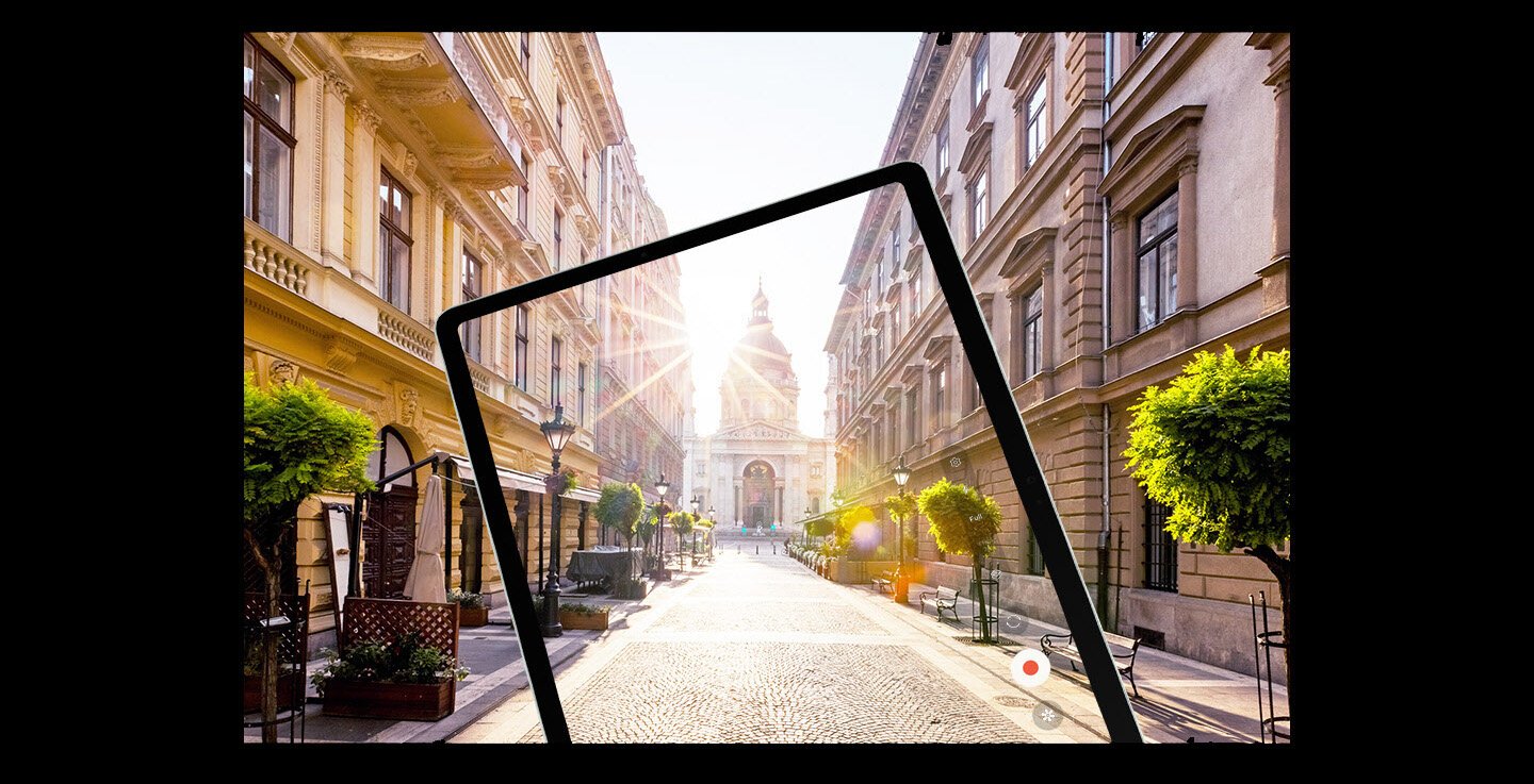 Tablet Samsung Galaxy Tab S9 FE w trybie nagrywania. Na ekranie widzimy ulicę z klasyczną europejską zabudową oraz flarę słońca padającego wprost w obiektyw. W tle przestrzeń miejska, która dopełnia widok wyświetlany na urządzeniu.