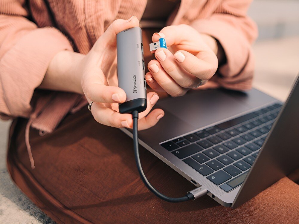Hub Verbatim 49147 Osoba w różowej kurtce trzyma hub USB podłączony do laptopa i pendrive gotowy do wpięcia, 
            hub obudowa aluminium wygląd kształt kompaktowy