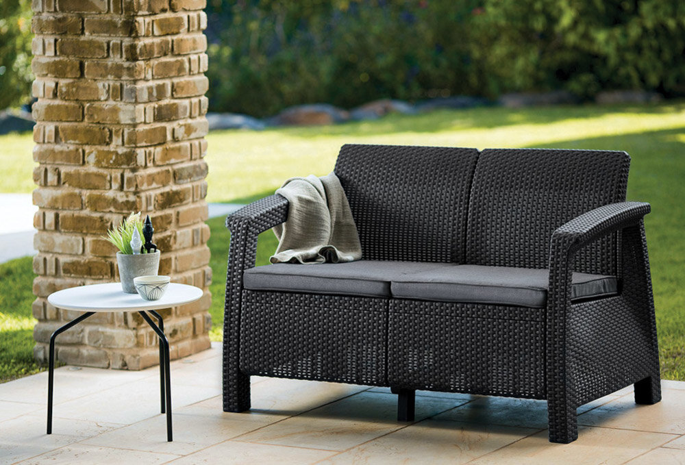 Sofa ogrodowa KETER Corfu II Love Brown 258946 Brąz sofa na tarasie w tle rośliny i drzewa z wysokiej jakości technorattanu odporny na uszkodzenia mechaniczne długa żywotność imitacja naturalnego splotu rattanowego sofa zachowa swój wygląd i funkcjonalność nie zblaknie nie popęka zabezpieczyć poduszki przed wilgocią i opadami