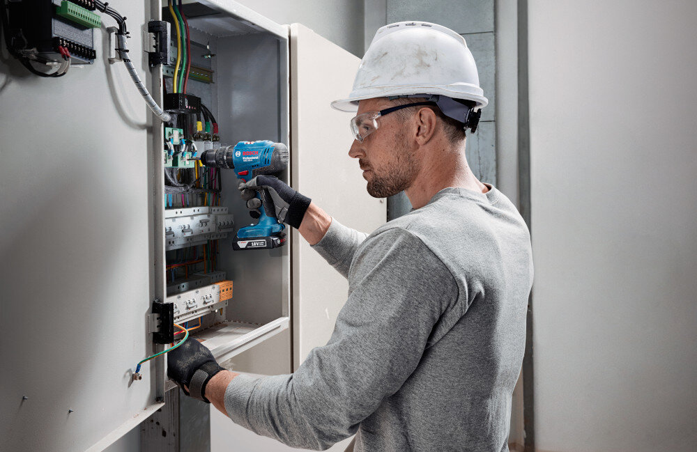 Wiertarko-wkrętarka BOSCH Professional GSB 183-LI 06019K9100 ściana skrzynka z czujnikami i kablami osoba używa wiertarko-wkrętarki wkręcanie wkrętów w drewnie i wiercenie w metalu i w murze funkcja udaru ułatwia organizację przestrzeni warsztatowej skraca czas realizacji projektów