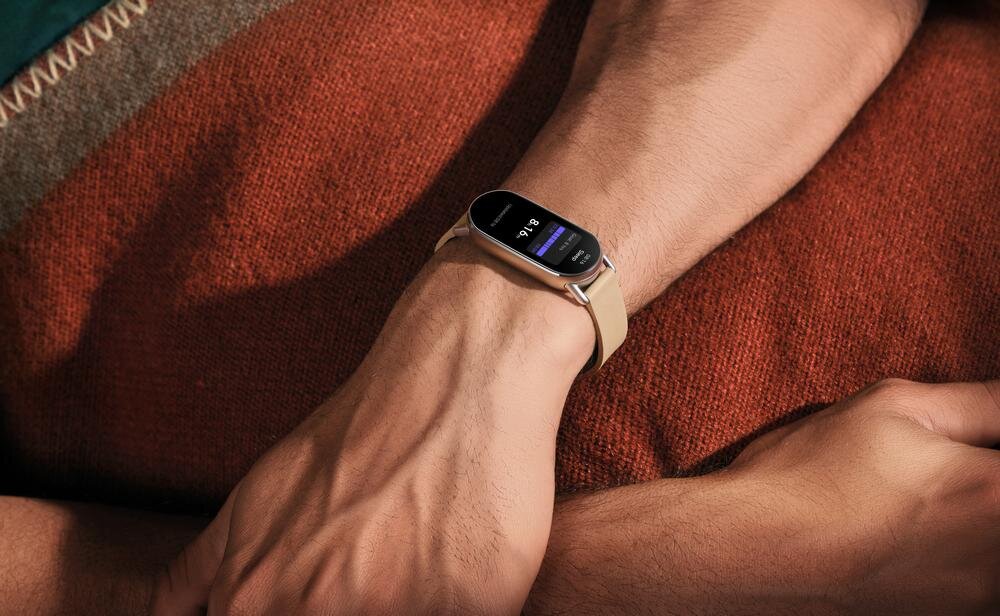 Na zdjęciu widoczny jest smartband z wyświetlaczem, na którym pokazana jest godzina 20:16. Opaska ma jasny pasek i jest noszona na nadgarstku osoby leżącej na czerwonej poduszce.