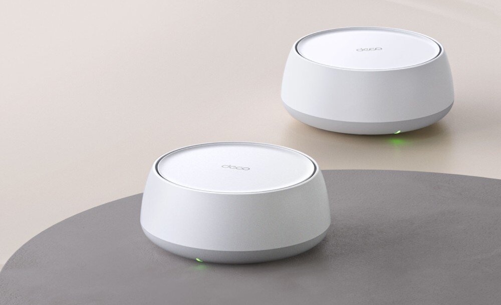 Router TP-LINK Deco BE25 Dwa routery o cylindrycznym kształcie w kolorze białym stojące na gładkiej, neutralnej powierzchni. Na dolnej krawędzi obu urządzeń świecą się zielone diody, opis
