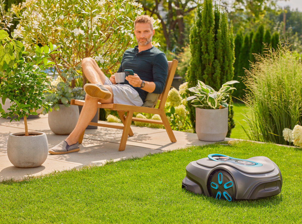 Robot koszący GARDENA Sielno Max 1200 19902-32 robot stoi na trawie w tle osoba siedzi na krześle na tarsie z telefonem w dłoni poziom hałasu 58 dB(A) akumulator litowo-jonowy o napięciu 18 V i pojemności 2,0 Ah dzienny czas pracy do 17 godzin