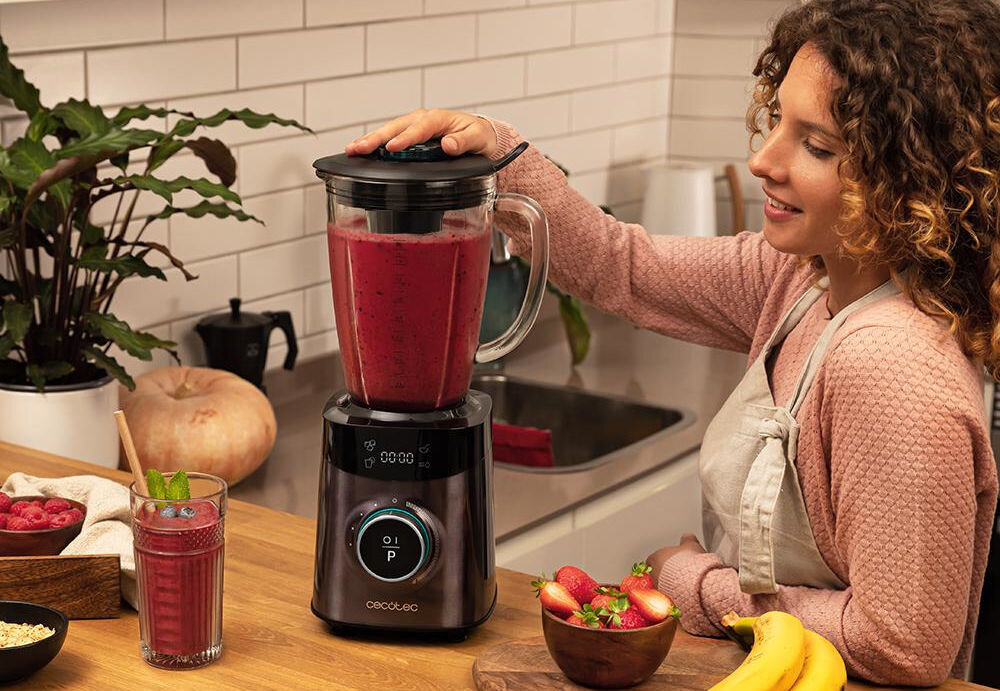 Blender kielichowy CECOTEC Power Black Titanium 2500 Advance blender stoi na blacie ciemna obudowa kobieta trzyma pokrywkę miksuje różowa masę pokrętło  36,5 x 21,7 x 27,7 cm 1800 W mocy aż 22 000 obrotów na minutę radzi sobie z twardymi składnikami orzechy nasiona kostki lodu potężna wydajność przygotowywanie wielu potraw szybkość precyzja 