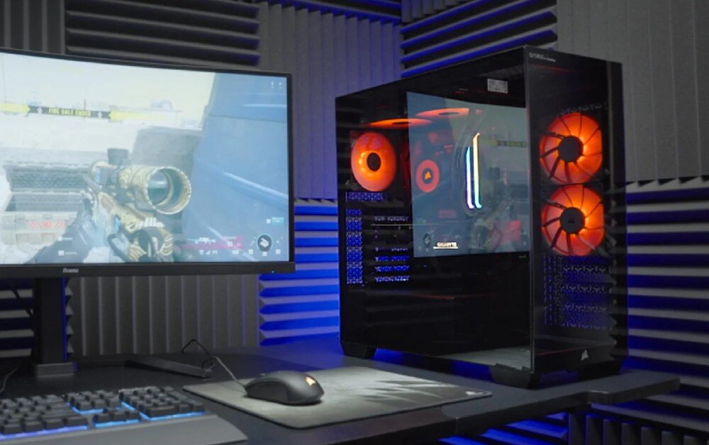 Komputer OPTIMUS E-sport GA550T-CR4 Komputer stacjonarny z przezroczystym panelem bocznym i czerwonym podświetleniem wnętrza stoi na biurku obok monitora, na którym trwa rozgrywka w grę FPS. W tle widać panele akustyczne na ścianie, opis