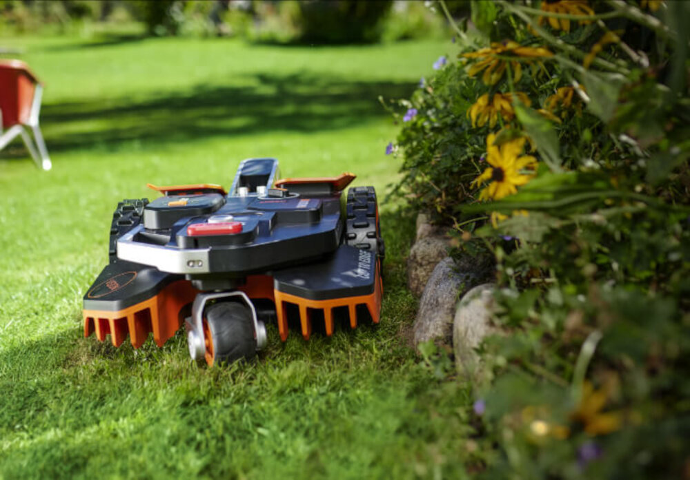 Robot koszący WORX Landroid Vision WR202E robot na trawie obok kamienie rabata kwiatowa automatyczne koszenie pielęgnacja trawnika łatwa obsługa aplikacja koszenie bez przewodów ograniczających