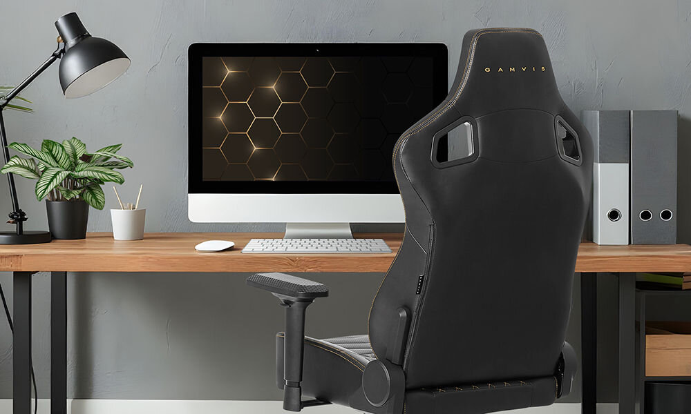 Fotel GAMVIS Elite 2.0 Pro XL Czarny (złote elementy) prezentacja fotela w pokoju przy biurku na nim monitor i akcesoria komputerowe kwiatek lampka brązowy blat biurka detale płaskie siedzisko ZeroStress+ dekoracyjne przeszycia welurowe poduszki wsparcie wygodnej pozycji